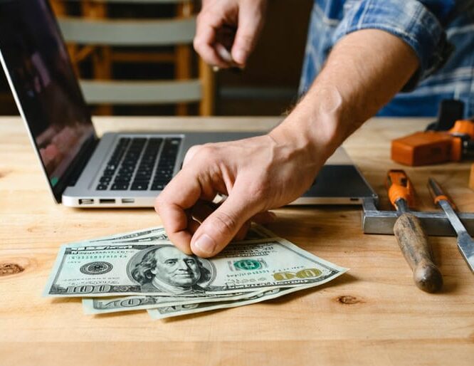 Woodworking student at a workbench with hand hovering over a small stack of cash and a debit card beside chisels and a measuring square, blurred laptop and half-built chair in a sunlit workshop background